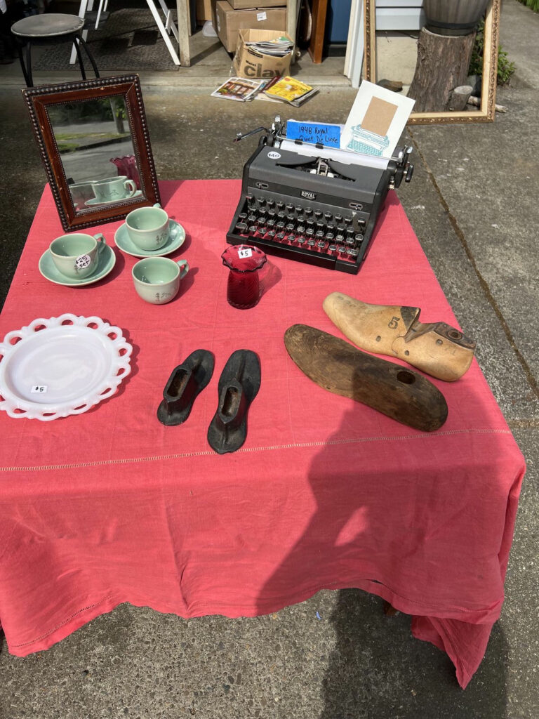 A red tablecloth-covered table at a yard sale displays vintage items, including a 1948 Royal Quiet De Luxe typewriter, green teacups and saucers, wooden shoe forms, and small decorative objects. A cardboard box labeled "clay" is visible in the background.