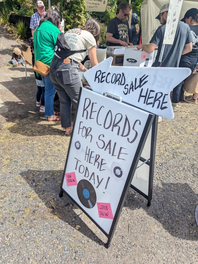 A crowd of people browses vinyl records at an outdoor record sale. A large white sign in the foreground reads “RECORDS FOR SALE HERE TODAY!” with hand-drawn records and pink notes saying “new stock in!” and “come on in!”