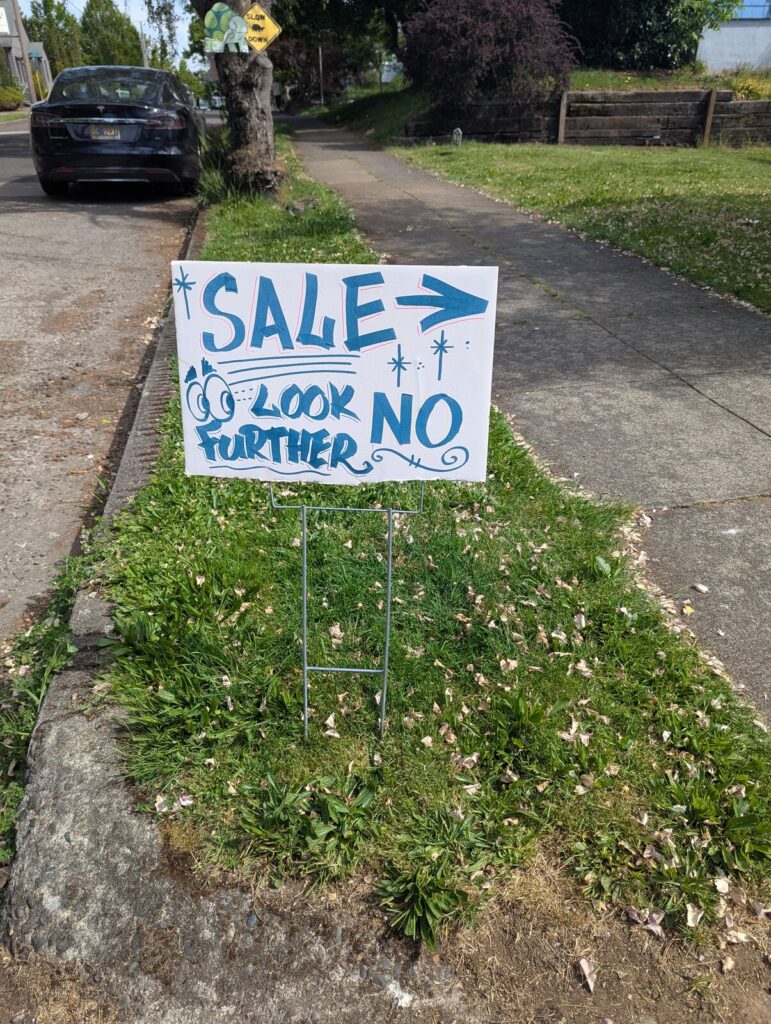 A handmade sign staked into a grassy curb reads “SALE → LOOK NO FURTHER” in bold blue letters with cartoon eyes and sparkles. A quiet residential sidewalk and parked car are in the background.