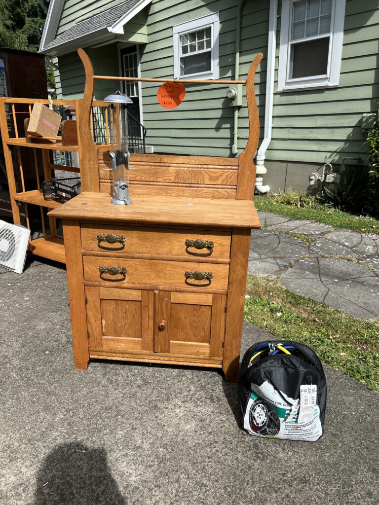 An antique wooden washstand with ornate drawer pulls is displayed on a driveway with a $100 price tag. A small bag labeled “FREE” containing tire chains sits next to it. A green house with white trim is visible behind it.